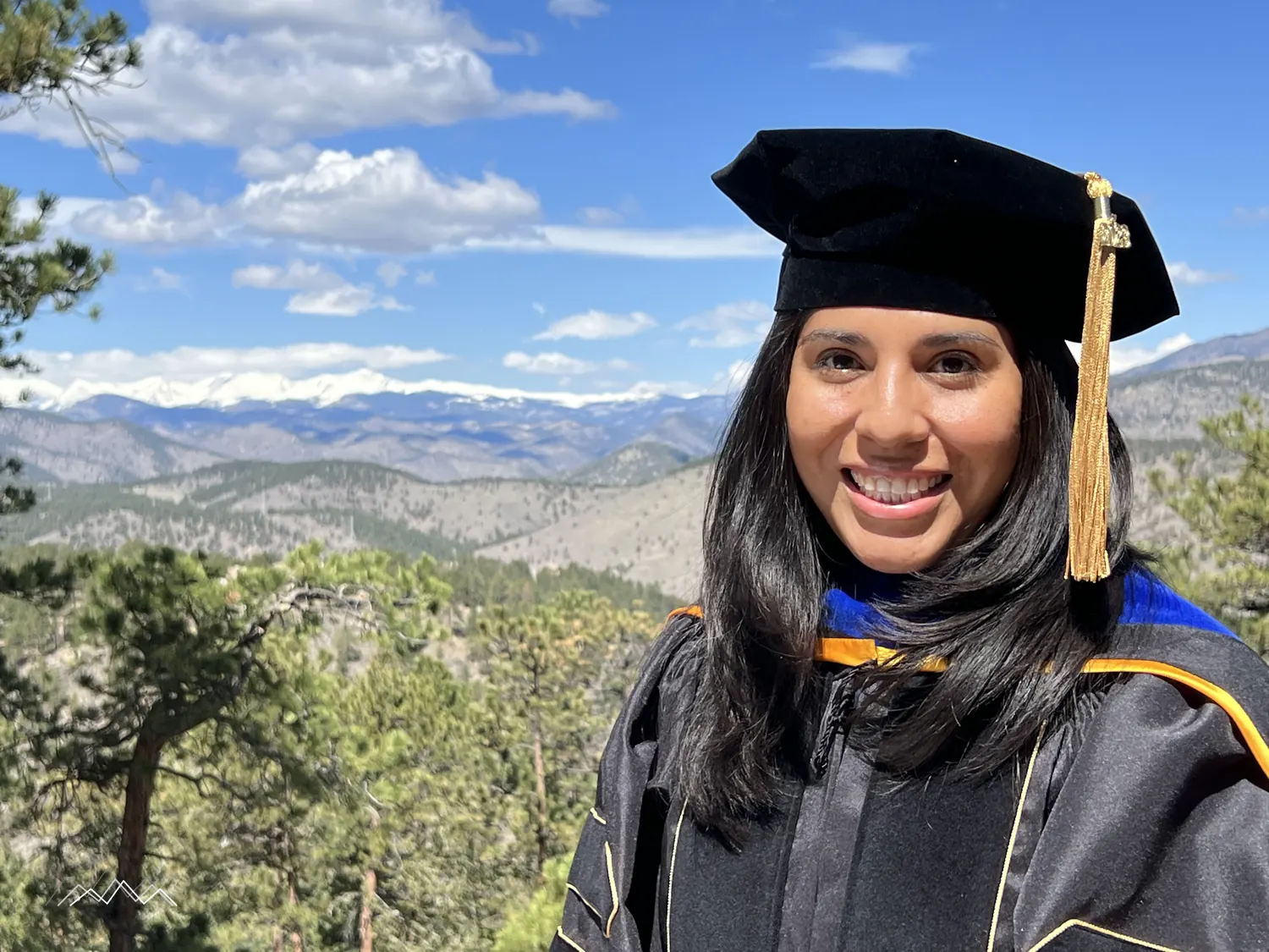 A doctoral graduate in cap and gown smiling in front of the snow-capped Indian Peaks