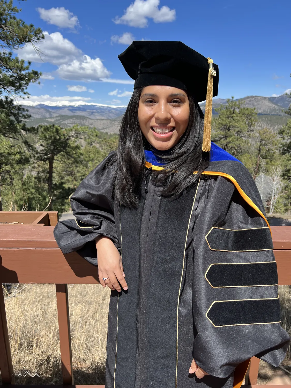A smiling doctoral graduate leaning on the deck railing, pine forest and snowy peaks behind