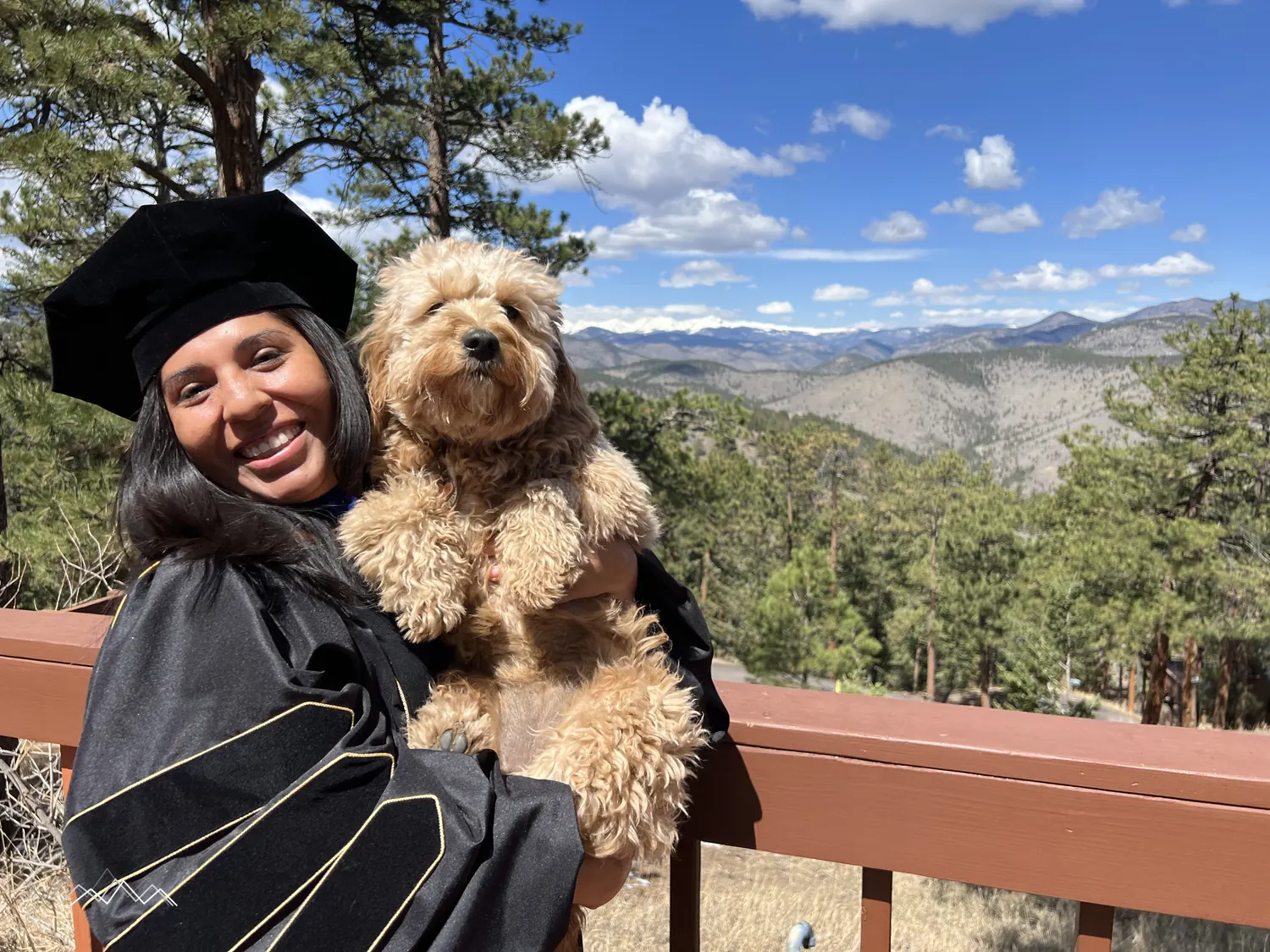 A doctoral graduate in cap and gown holding a fluffy golden dog on the deck, mountains behind