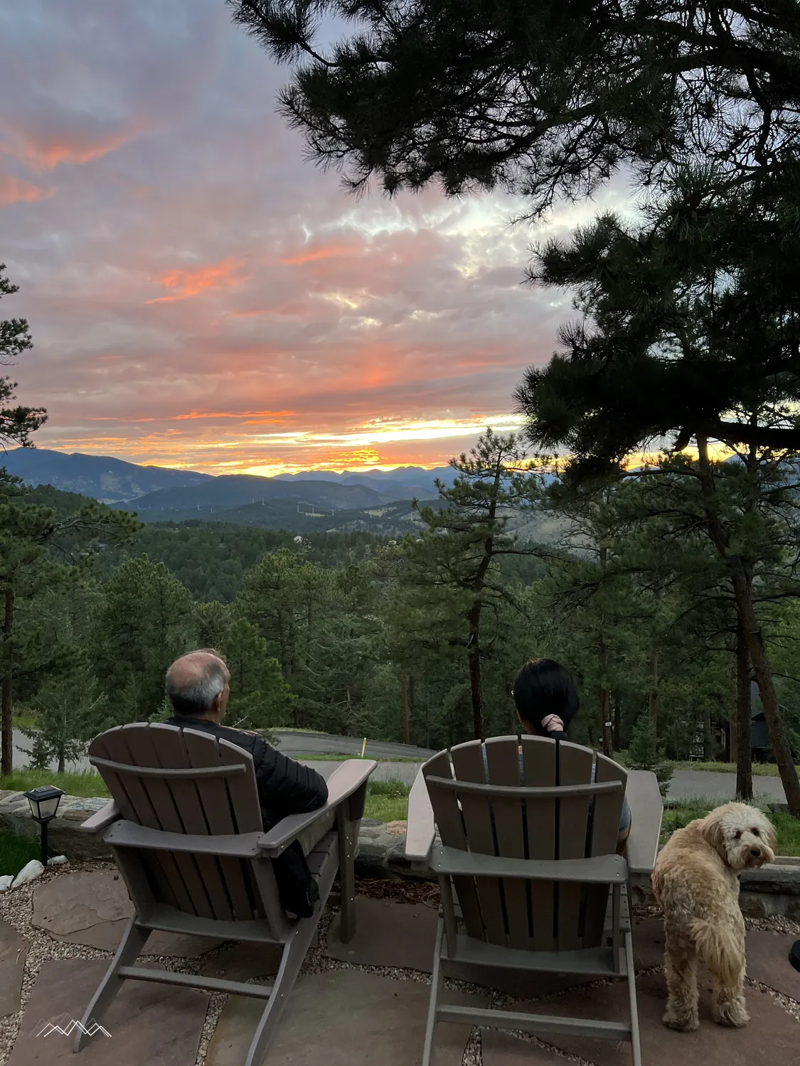 Two people relaxing in Adirondack chairs watching a pink sunset over the mountains, a golden dog looking back at the camera