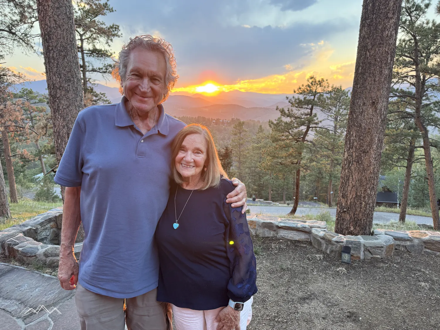 An older couple smiling together on the patio at sunset, the sun setting over the mountains between the pines