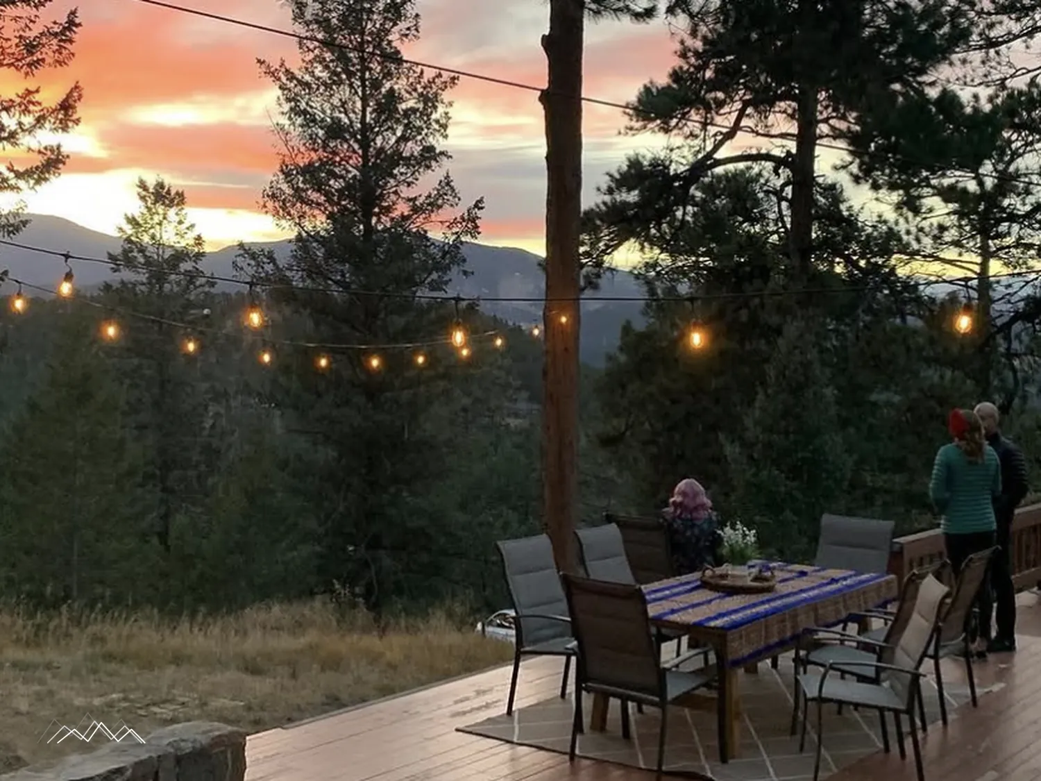 A dinner table set on the deck under glowing string lights, with a pink sunset over the mountains