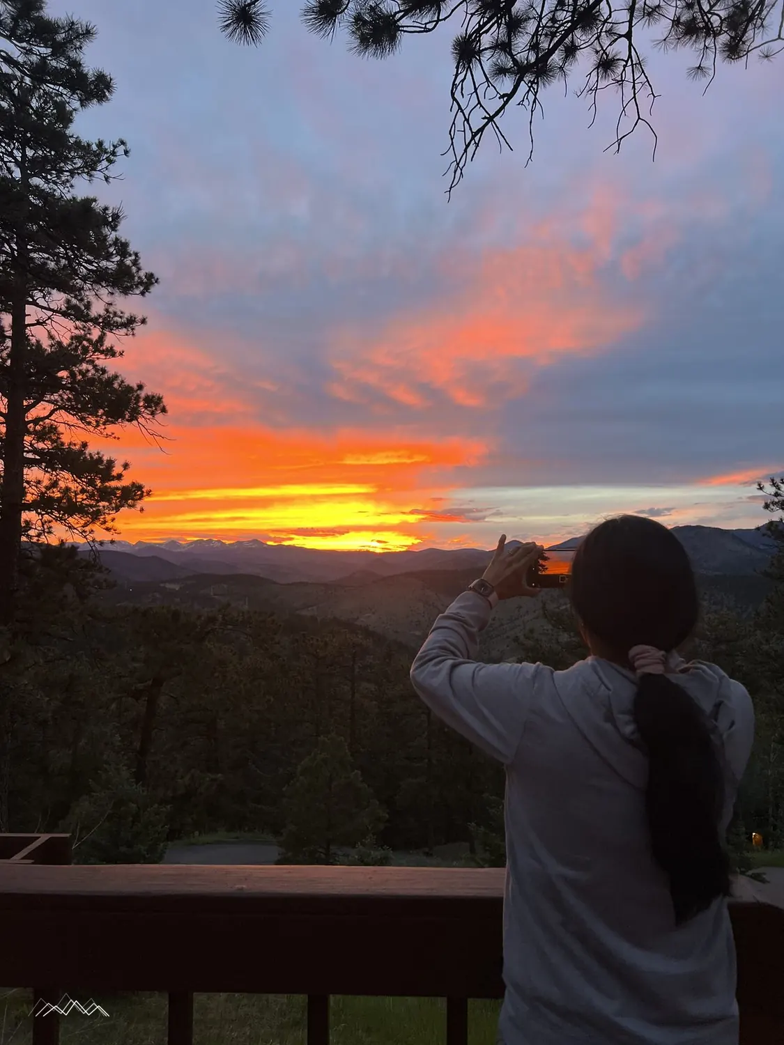Someone at the deck railing taking a photo of a fiery pink and orange sunset over the mountains