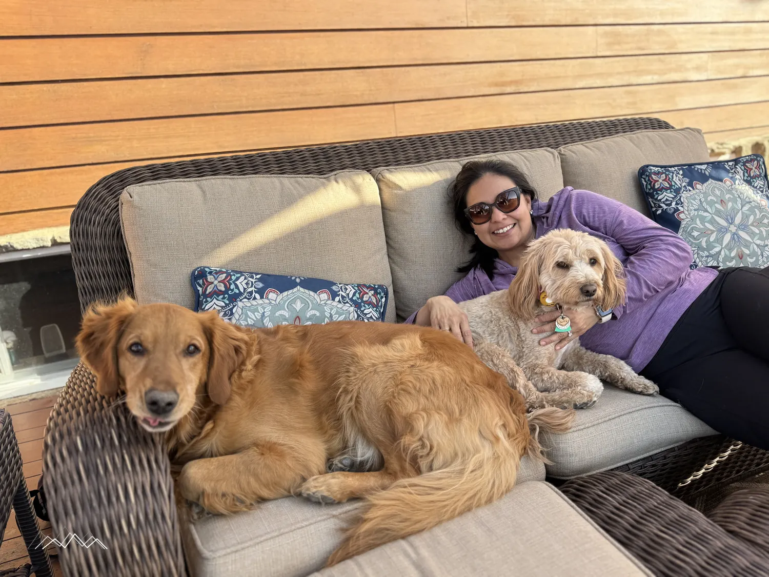 A smiling woman relaxing on an outdoor wicker couch with Maisie the golden retriever and Bella the golden doodle