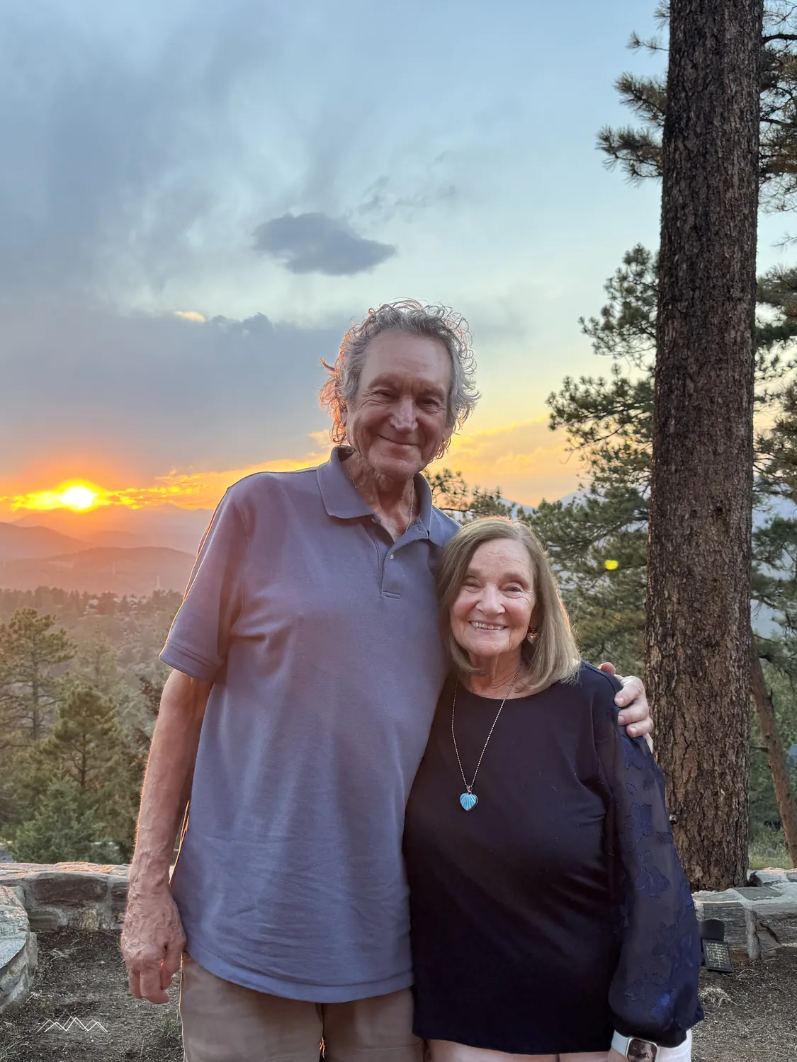 An older couple smiling together by the stone wall at sunset, the sun setting over the mountains between the pines