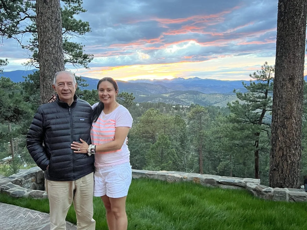 Mily und ihr Vater lächeln gemeinsam auf dem Patio bei Sonnenuntergang, dahinter rosa Wolken über den Bergen und Kiefern