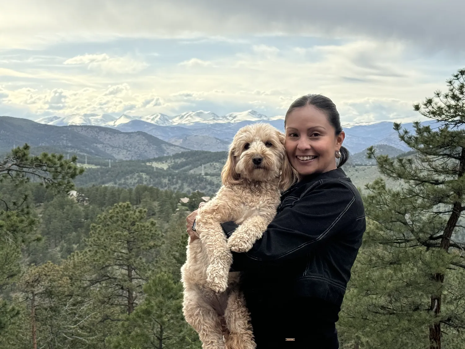 Mily souriante tenant Bella, la golden doodle, avec les sommets enneigés et la forêt de pins derrière